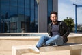 Handsome young man with beard, sunglasses, leather jacket and jeans, sitting on a stone bench, relaxed. Concept beauty, fashion, Royalty Free Stock Photo