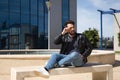 Handsome young man with beard, sunglasses, leather jacket and jeans, sitting on a stone bench, relaxed. Concept beauty, fashion, Royalty Free Stock Photo