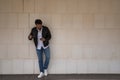 Handsome young man with beard, sunglasses, leather jacket, white shirt and jeans, leaning against a white wall, consulting his Royalty Free Stock Photo