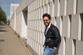 Handsome young man with beard, sunglasses, leather jacket, white shirt and jeans, leaning against a wall smiling very happy. Royalty Free Stock Photo