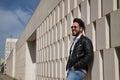 Handsome young man with beard, sunglasses, leather jacket, white shirt and jeans, leaning against a wall smiling very happy. Royalty Free Stock Photo