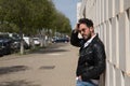 Handsome young man with beard, sunglasses, leather jacket, white shirt and jeans, leaning against a wall of white stone blocks, Royalty Free Stock Photo