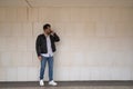 Handsome young man with beard, sunglasses, leather jacket, white shirt and jeans, next to a white wall, talking on his cell phone Royalty Free Stock Photo