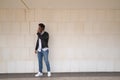 Handsome young man with beard, sunglasses, leather jacket, white shirt and jeans, next to a white wall, talking on his cell phone Royalty Free Stock Photo