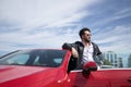 Handsome young man with beard, sunglasses, leather jacket and white shirt, leaning on the roof of his red sports car. Concept