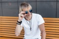 Handsome young man blond with stylish hairstyle in a trendy t-shirt in vintage round sunglasses is resting sitting on a wooden Royalty Free Stock Photo