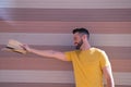 A handsome young man with blue eyes and a beard and a yellow T-shirt stretches out his arm with a hat in his hand. In the Royalty Free Stock Photo