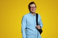 Handsome young man in blue shirt and eyeglasses holding backpack and smiling at camera on yellow backdrop Royalty Free Stock Photo