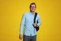 Handsome young man in blue shirt and eyeglasses holding backpack and smiling at camera on yellow backdrop Royalty Free Stock Photo