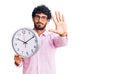 Handsome young man with curly hair and bear holding big clock with open hand doing stop sign with serious and confident Royalty Free Stock Photo