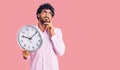 Handsome young man with curly hair and bear holding big clock serious face thinking about question with hand on chin, thoughtful Royalty Free Stock Photo