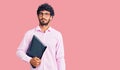 Handsome young man with curly hair and bear holding business folder thinking attitude and sober expression looking self confident Royalty Free Stock Photo