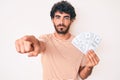 Handsome young man with curly hair and bear holding money bag with dollar symbol pointing with finger to the camera and to you, Royalty Free Stock Photo