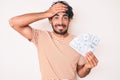 Handsome young man with curly hair and bear holding money bag with dollar symbol stressed and frustrated with hand on head, Royalty Free Stock Photo