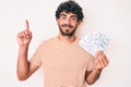 Handsome young man with curly hair and bear holding money bag with dollar symbol surprised with an idea or question pointing Royalty Free Stock Photo