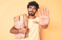 Handsome young man with curly hair and bear holding paper bag with bread with open hand doing stop sign with serious and confident Royalty Free Stock Photo