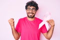 Handsome young man with curly hair and bear holding pink cancer ribbon screaming proud, celebrating victory and success very Royalty Free Stock Photo