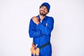 Handsome young man with curly hair and bear weaing handyman uniform hugging oneself happy and positive, smiling confident Royalty Free Stock Photo