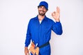 Handsome young man with curly hair and bear weaing handyman uniform smiling positive doing ok sign with hand and fingers Royalty Free Stock Photo