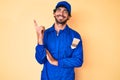 Handsome young man with curly hair and bear wearing builder jumpsuit uniform with a big smile on face, pointing with hand and Royalty Free Stock Photo