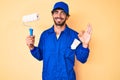 Handsome young man with curly hair and bear wearing builder jumpsuit uniform holding paint roller doing ok sign with fingers, Royalty Free Stock Photo