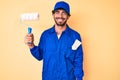 Handsome young man with curly hair and bear wearing builder jumpsuit uniform holding paint roller looking positive and happy Royalty Free Stock Photo