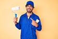 Handsome young man with curly hair and bear wearing builder jumpsuit uniform holding paint roller smiling happy pointing with hand Royalty Free Stock Photo