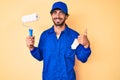 Handsome young man with curly hair and bear wearing builder jumpsuit uniform holding paint roller smiling happy and positive, Royalty Free Stock Photo