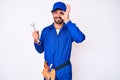 Handsome young man with curly hair and bear wearing builder jumpsuit uniform and holding wrench smiling happy doing ok sign with Royalty Free Stock Photo