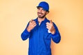 Handsome young man with curly hair and bear wearing builder jumpsuit uniform pointing fingers to camera with happy and funny face Royalty Free Stock Photo