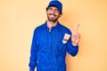 Handsome young man with curly hair and bear wearing builder jumpsuit uniform smiling with happy face winking at the camera doing Royalty Free Stock Photo