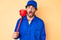 Handsome young man with curly hair and bear wearing plumber uniform holding toilet plunger scared and amazed with open mouth for