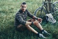 Handsome Young Man Cyclist Sits On Summer Meadow Near Bicycle, Holding At Tablet Recreation Resting Travel Destination Concept