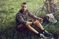 Handsome Young Man Cyclist Sits On Summer Meadow Near Bicycle, Holding At Tablet Recreation Resting Travel Destination Concept