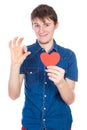 Handsome young man in denim blue shirt standing on a white background with a red paper heart in hands.