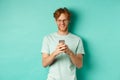 Handsome young man in glasses with red messy hair reading message on mobile phone, smiling and looking at screen Royalty Free Stock Photo