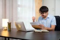 A handsome young man in glasses showing success gesture while reading acceptance letter with laptop on desk at home Royalty Free Stock Photo