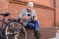 Handsome young man in grey coat and hat talking on the mobile phone and smiling while sitting near his bicycle outdoors Royalty Free Stock Photo