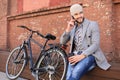 Handsome young man in grey coat and hat talking on the mobile phone and smiling while sitting near his bicycle outdoors Royalty Free Stock Photo