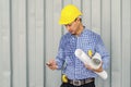 Handsome young man in hardhat holding blueprint and using mobile phone while standing outdoors and against building structure Royalty Free Stock Photo