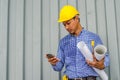 Handsome young man in hardhat holding blueprint and using mobile phone while standing outdoors and against building structure Royalty Free Stock Photo