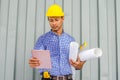 Handsome young man in hardhat holding blueprint and using tablet while standing outdoors and against building structure Royalty Free Stock Photo