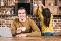 handsome young man holding cup of coffee and smiling at camera while using laptop and girlfriend Royalty Free Stock Photo