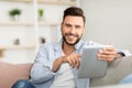 Handsome young man holding digital tablet and smiling at camera, sitting on couch at home, free space Royalty Free Stock Photo