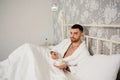 A handsome young man is lying in bed in a white Terry robe and drinking a morning drink Royalty Free Stock Photo