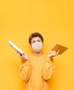 Handsome young man in a medical mask isolated with a book and notebook on a yellow background, looking at the camera and puzzled. Royalty Free Stock Photo