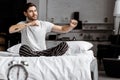 Handsome young man in pajamas stretching hands while sitting on bed and looking Royalty Free Stock Photo