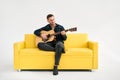 Handsome young man playing acoustic guitar whlile sitting on yellow sofa over white background Royalty Free Stock Photo