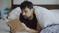 Handsome young man reading a book lying in bed under blanket at home Royalty Free Stock Photo