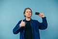 Handsome young man in shirt with bank card in hand, showing thumbs up and smiling looking at camera on blue background Royalty Free Stock Photo